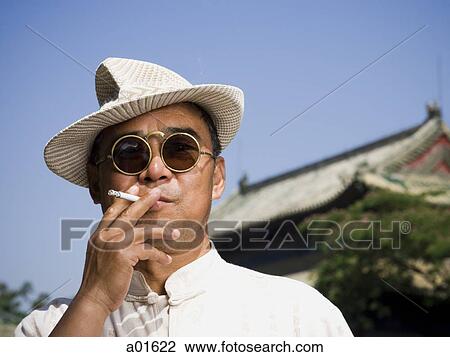 Stock Image - Man with sunglasses and straw hat smoking cigarette outdoors with pagoda in background. Fotosearch