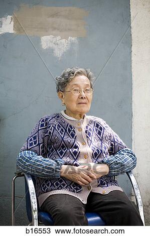 Stock Image - Mature woman with eyeglasses sitting in blue chair. Fotosearch