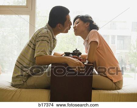 Picture - Profile of couple sitting cross legged kissing with tea and fruit with large window in background. Fotosearch