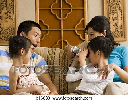 Stock Image - Son recording father and sister with video camera and mother on sofa. Fotosearch