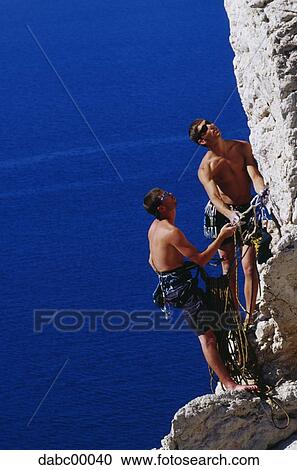 Stock Image - Two men climbing mountain with harnesses and ropes. Fotosearch