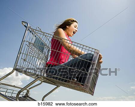 Stock Image - Woman in shopping cart outdoors with arms up smiling. Fotosearch