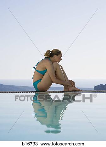 Stock Image - Woman in swimsuit sitting at edge of infinity pool outdoors. Fotosearch
