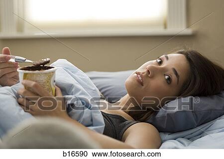 Stock Image - Woman lying in bed eating chocolate ice cream. Fotosearch
