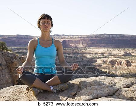 Stock Image - Woman sitting cross legged on rock outdoors doing yoga. Fotosearch
