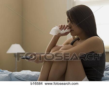 Picture - Woman sitting on bed with television remote and tissue. Fotosearch