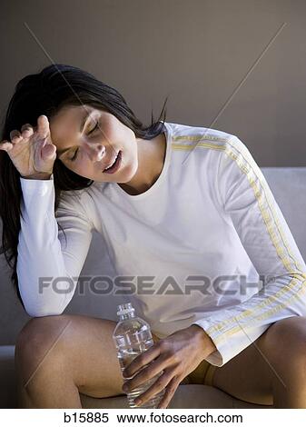 Woman sitting on sofa with bottled water wiping brow View Large Photo Image Stock Photography - Woman sitting on sofa with bottled water wiping brow. Fotosearch