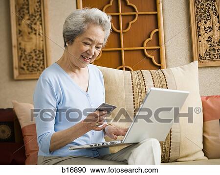 Stock Image - Woman sitting on sofa with laptop and credit card smiling. Fotosearch