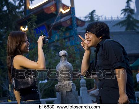 Stock Image - Woman taking photo outdoors of man making peace sign and smiling. Fotosearch