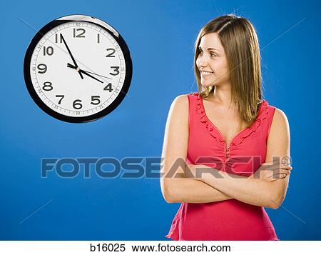 Stock Photography - Woman with arms crossed looking at wall clock attached to plumbing pipes. Fotosearch