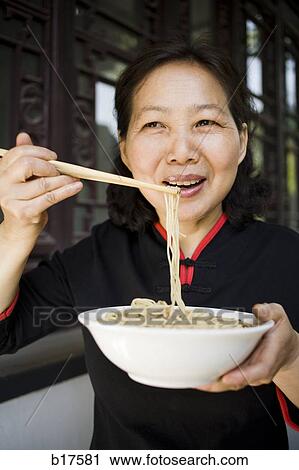 Stock Image - Woman with bowl of noodles and chopsticks eating. Fotosearch