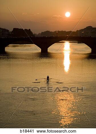Man rowing on a body of water with a bridge and setting sun in the background. View Large Photo Image Stock Image - Man rowing on a body of water with a bridge and setting sun in the background.. Fotosearch