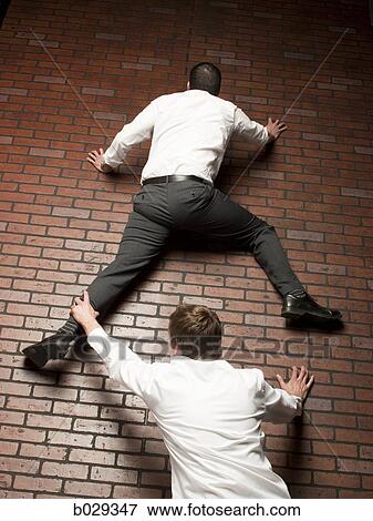 Stock Photo - man climbing up a brick wall while another man grabs on to his leg. Fotosearch