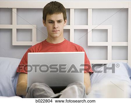Picture - man working on a laptop on a bed. Fotosearch