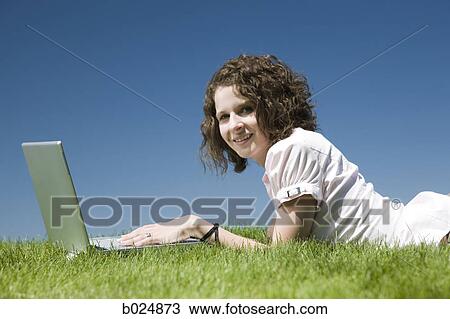 Stock Image - woman lying on the grass using a laptop. Fotosearch
