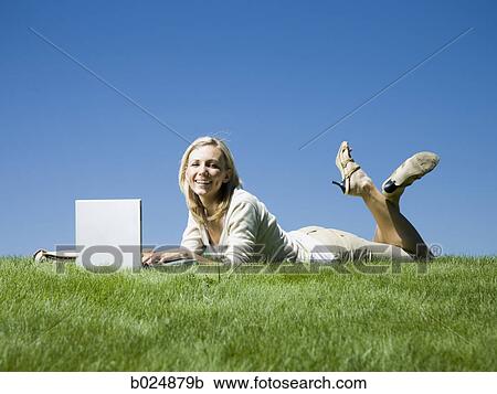 Stock Image - woman lying on the grass using a laptop. Fotosearch