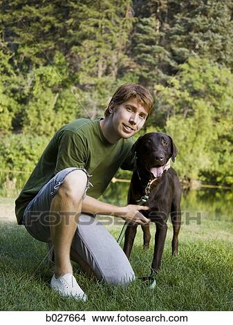 Picture - young man kneeling with a chocolate labrador retriever. Fotosearch