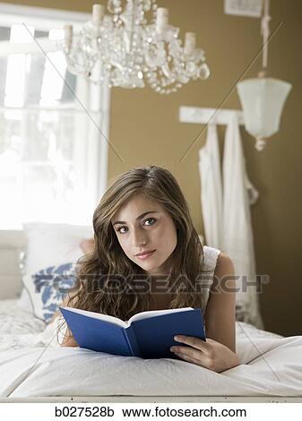 Stock Image - young woman lying on her bed reading a book. Fotosearch