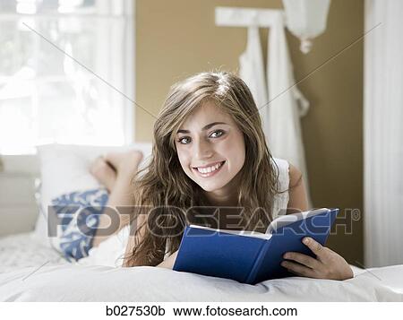 Stock Image - young woman lying on her bed reading a book. Fotosearch