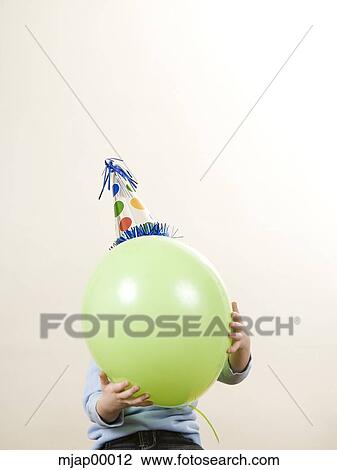 child wearing a party hat holding a balloon in front of his face View Large Photo Image Stock Image - child wearing a party hat holding a balloon in front of his face. Fotosearch
