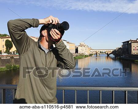 Stock Image - Italy, Florence, Man using camera on bridge over River Arno . Fotosearch
