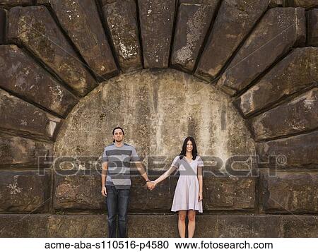 Stock Image - Italy, Florence, Portrait of young couple holding hands by stone wall. Fotosearch