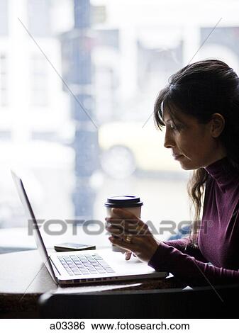 Stock Photograph - One mid adult woman drinking coffee next to laptop in cafe. Fotosearch