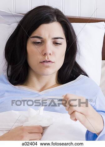 Picture - Orem, Utah, USA, young ill woman in bed looking at thermometer.. Fotosearch