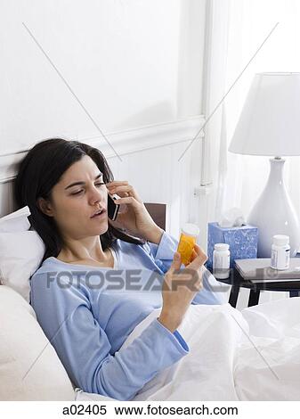 Stock Photography - Orem, Utah, USA, young ill woman in bed looking at pill bottle and talking on the phone. Fotosearch
