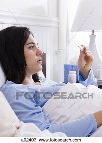 Stock Image - Orem, Utah, USA, young ill woman l in bed looking at thermometer. Fotosearch