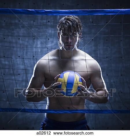 Stock Image - Studio shot of volleyball player standing behind net and holding ball. Fotosearch