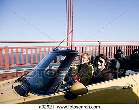 Stock Image - USA, California, San Francisco, young couple passing Golden Gate Bridge in convertible car. Fotosearch