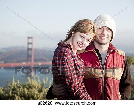 Stock Photo - USA, California, San Francisco, young couple standing, Golden Gate Bridge in background. Fotosearch