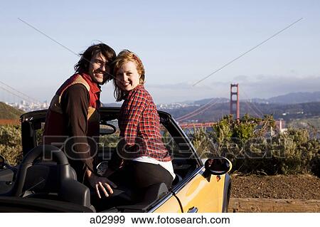 Stock Photo - USA, California, San Francisco, young couple sitting car with Golden Gate Bridge in background, portrait. Fotosearch