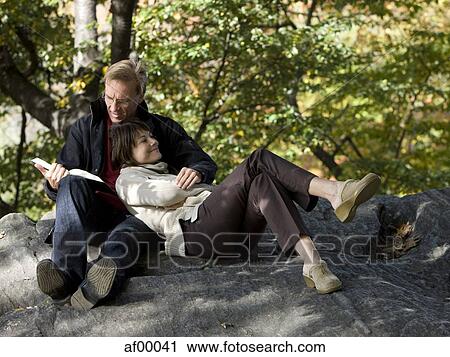 Stock Image - USA, New York City, Manhattan, Central Park, Mature couple reading book in Central Park. Fotosearch