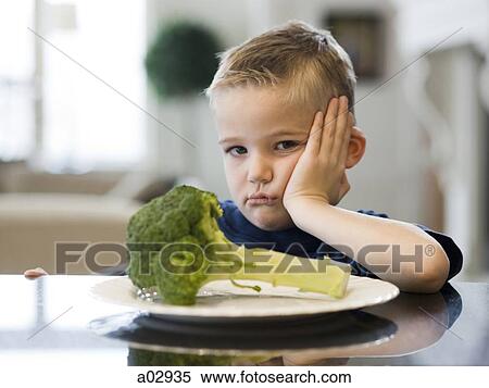 Stock Photography - USA, Utah, Alpine, boy (6-7) sulking by table with plate of broccoli. Fotosearch