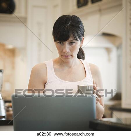 Stock Image - USA, Utah, Alpine, mid adult woman standing near laptop and holding mug. Fotosearch