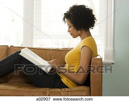Stock Photo - USA, Utah, Provo, young woman reading book on couch, side view. Fotosearch