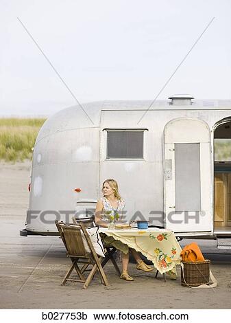 Stock Image - woman sitting at a table on the beach outside her airstream classic trailer. Fotosearch