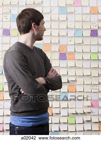 Stock Image - Young man standing in front of wall covered with adhesive notes and reading. Fotosearch