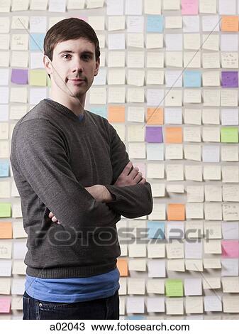 Stock Image - Young man standing in front of wall covered with adhesive notes, portrait. Fotosearch