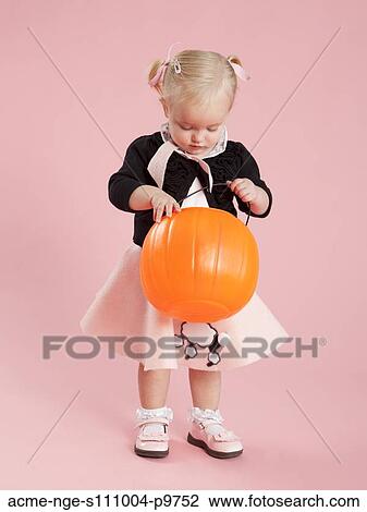 Stock Image - Baby girl (12-17 months) in 1950s style costume, holding pumpkin lantern for Halloween. Fotosearch
