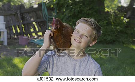 Stock Image - High angle portrait of smiling boy petting rooster on shoulder in backyard. Fotosearch
