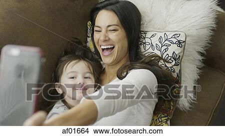 High angle selective focus view of playful mother and daughter posing and taking selfie with smartphone on sofa View Large Photo Image Picture - High angle selective focus view of playful mother and daughter posing and taking selfie with smartphone on sofa. Fotosearch