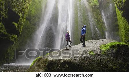 Low angle view of couple standing on mossy boulder admiring waterfall in remote landscape View Large Photo Image Stock Image - Low angle view of couple standing on mossy boulder admiring waterfall in remote landscape. Fotosearch