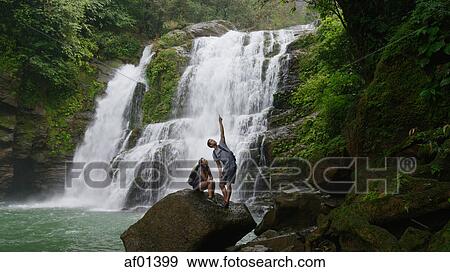 Stock Photo - Low angle view of couple standing on boulder pointing and admiring scenic view near tropical waterfall. Fotosearch