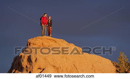 Stock Photo - Low angle view of couple standing on remote boulder under sunset sky. Fotosearch