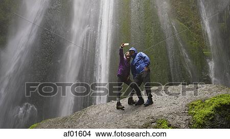 Low angle view of couple taking smartphone selfie standing on mossy boulder near waterfall in remote landscape View Large Photo Image Picture - Low angle view of couple taking smartphone selfie standing on mossy boulder near waterfall in remote landscape. Fotosearch