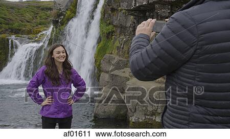 Man photographing smiling girlfriend posing near waterfall over remote rock cliffs View Large Photo Image Stock Image - Man photographing smiling girlfriend posing near waterfall over remote rock cliffs. Fotosearch