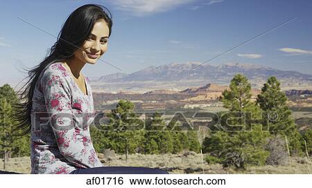 Portrait of smiling woman sitting near scenic view of mountains, trees and scrubland in remote landscape under blue sky View Large Photo Image Stock Photograph - Portrait of smiling woman sitting near scenic view of mountains, trees and scrubland in remote landscape under blue sky. Fotosearch